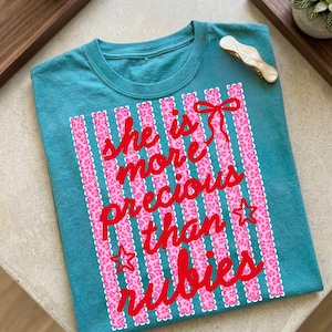 Può includere: T-shirt turchese con la scritta rossa "She is more precious than rubies". Il design include strisce verticali rosa con motivo leopardato, un fiocco rosso e stelle.