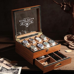 May include: A wooden watch box with a hinged lid, displaying several wristwatches. The box has a drawer and is lined with a light-colored material. The lid has the text "RAVEN SMITH" and a logo. The box is on a wooden surface.