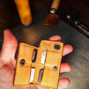 Può includere: Un piccolo porta SD in pelle marrone chiaro con cuciture gialle. Il porta SD è aperto, rivelando diverse schede SD. È tenuto in mano, con strumenti e filo sullo sfondo.