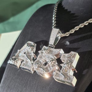 May include: A silver-toned pendant necklace featuring the numbers 451 in a block font. The surface of the numbers is covered in small, clear, sparkling stones and engraved rose details. The pendant hangs from a thick, twisted silver-toned rope chain.
