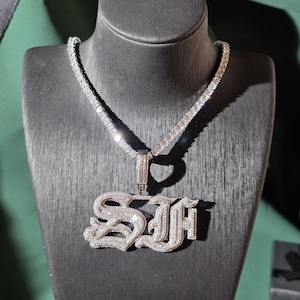 May include: A silver-toned necklace featuring a tennis chain and a large, ornate pendant shaped like the letters SJ. The entire surface of the pendant and the chain links are encrusted with numerous small, clear, reflective stones.