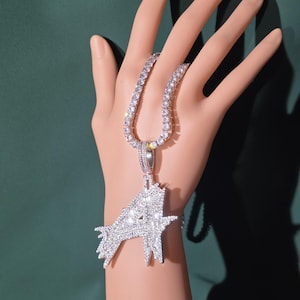 May include: A silver-toned necklace with a large, sparkling pendant in the shape of the letter "A". The necklace features a tennis chain design with numerous small, clear stones. The pendant is encrusted with many clear, sparkling stones.