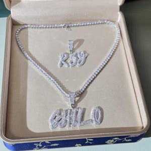 May include: A diamond-encrusted necklace and pendant set in a presentation box. The necklace is a tennis style, and the pendant spells out "CHILLO". A smaller pendant above spells "Roy". The box is lined with a light beige fabric and has a blue floral design.
