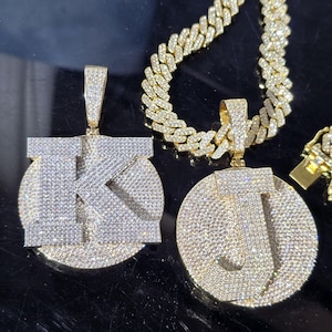 May include: Two gold-toned circular pendants featuring the letters K and J, both encrusted with numerous small clear stones. The pendants are attached to a thick, gold-toned cuban link chain that is also covered in small, sparkling clear stones.