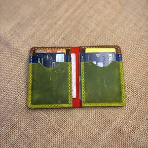 Può includere: Portafoglio in pelle aperto con più fessure per carte. Il portafoglio è aperto, rivelando diverse carte di credito e un interno rosso. L'esterno presenta fessure verdi e cuciture gialle. Il portafoglio è su una superficie beige strutturata.