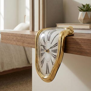 Puede incluir: Un reloj dorado y derretido con una esfera blanca y números romanos negros. El reloj descansa sobre una estantería de madera, con libros y una pequeña planta en maceta en el fondo. El diseño del reloj es surrealista.
