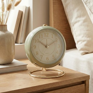 May include: A round, sage green quartz clock with a gold-colored base. The clock face is white with black numerals and gold hands. The clock is sitting on a wooden nightstand.