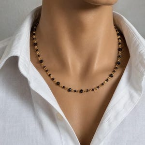 Puede incluir: Un collar de cadena dorada con pequeñas cuentas negras facetadas espaciadas uniformemente. El collar descansa sobre el cuello, mostrando un diseño de eslabones delicados que alterna entre segmentos de metal dorado pulido y cuentas oscuras.