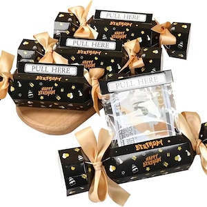 May include: Black birthday crackers with gold bows and the text "PULL HERE" and "HAPPY BIRTHDAY". The crackers are decorated with cake and star graphics. Some crackers are open, revealing contents.