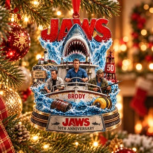 Könnte beinhalten: Ein Jaws 50th Anniversary Weihnachtsanhänger mit einem Hai, der hinter einem Boot mit drei Figuren aus blauem Wasser auftaucht. Das Boot ist mit Brody beschriftet und ein Schild zeigt Amity Island. Der Sockel trägt das Jaws-Logo und 50th Anniversary.