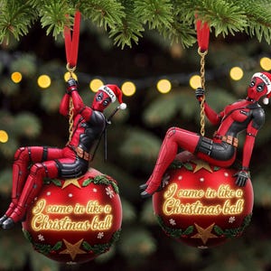 Pode incluir: Dois enfeites de Natal do personagem Deadpool em vermelho e preto. Cada figura usa um gorro de Papai Noel com um nome, Robert ou Ashley, e está sentada sobre uma bola vermelha com estrelas douradas, azevinho verde e o texto I came in like a Christmas ball.