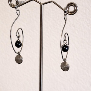 May include: Silver-toned earrings with a unique spiral design. Each earring features a small black bead and a circular pendant with a spiral pattern. The earrings are displayed on a silver metal stand.