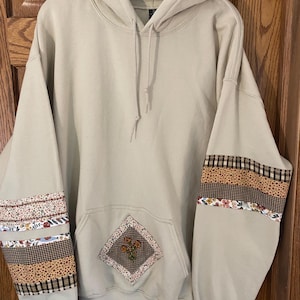 May include: A beige hooded sweatshirt featuring long sleeves with horizontal bands of brown plaid, yellow floral, and white floral fabric patches. A diamond-shaped patch with a brown checkered border and a small floral embroidery sits on the front pocket.