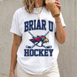 May include: White t-shirt with the text "BRIAR U HOCKEY" in navy blue block letters. The shirt features a logo of a stylized bird with crossed hockey sticks. The bird is red, white, and blue. The shirt is worn with white shorts.