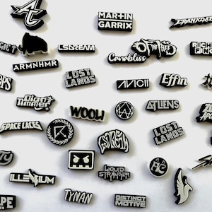 May include: A collection of black and white shoe charms featuring various music artist logos and festival names including Martin Garrix, Avicii, Lost Lands, Wooli, Space Laces, Levity, Illenium, Escape Halloween, and others in diverse fonts and shapes.
