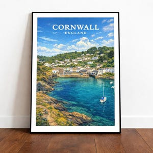 May include: A framed print featuring a coastal scene of Cornwall, England. The image showcases a vibrant blue bay with a sailboat, surrounded by rocky cliffs and a village. The sky is a bright blue with fluffy clouds. The print is displayed on a wooden floor.