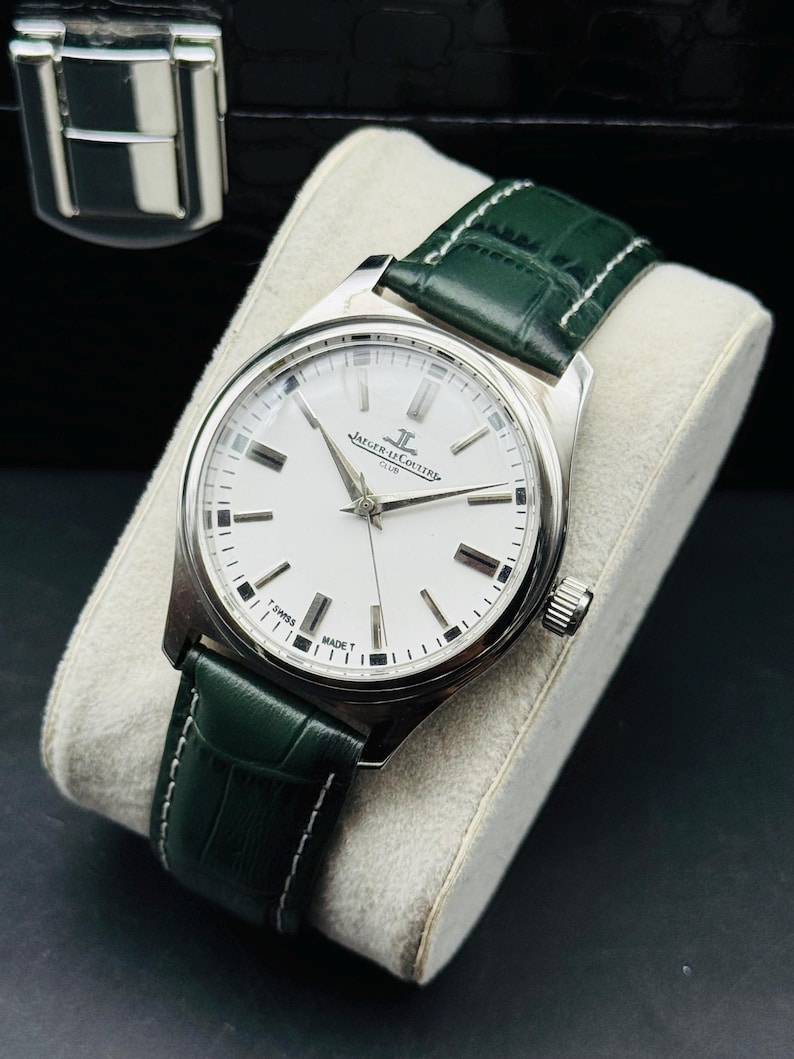 May include: A silver-toned Jaeger-LeCoultre Club watch with a white face and green leather strap. The watch has a round face with black markers and hands. The green strap is stitched with white thread. The watch is displayed on a white cushion.