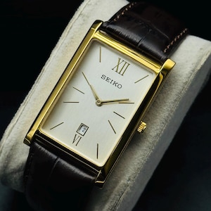 Könnte beinhalten: Eine rechteckige Seiko-Uhr mit goldfarbenem Gehäuse und cremefarbenem Zifferblatt. Die Uhr hat goldene Zeiger und Stundenmarkierungen, mit der römischen Ziffer XII oben. Das braune Lederarmband wird mit einer Schnalle befestigt. Die Uhr wird auf einem grauen Kissen präsentiert.