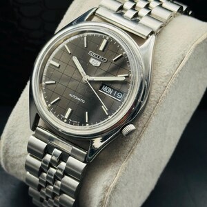 Op de afbeelding: Een zilverkleurig Seiko 5 automatisch horloge met een donkergrijze geruite wijzerplaat. Het horloge heeft een datumweergave met "MON 19" en een zilveren armband. Het horloge wordt getoond op een lichtgrijs kussen.