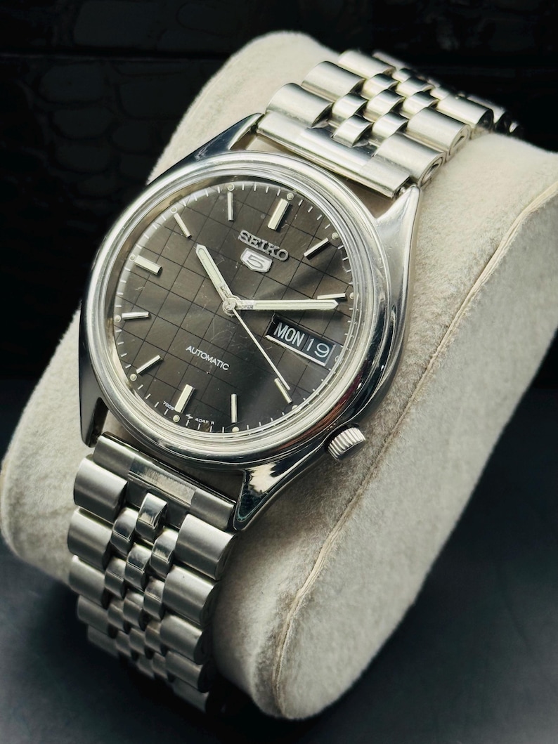 Op de afbeelding: Een zilverkleurig Seiko 5 automatisch horloge met een donkergrijze geruite wijzerplaat. Het horloge heeft een datumweergave met "MON 19" en een zilveren armband. Het horloge wordt getoond op een lichtgrijs kussen.