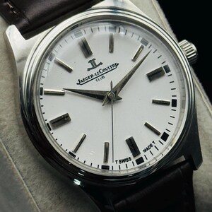 May include: A silver-toned Jaeger-LeCoultre Club watch with a white face and black markers. The watch has a dark brown leather band. The watch face displays the brand name and "MADE T SWISS".