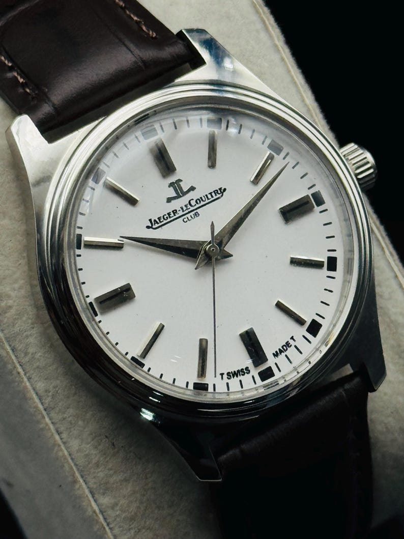 May include: A silver-toned Jaeger-LeCoultre Club watch with a white face and black markers. The watch has a dark brown leather band. The watch face displays the brand name and "MADE T SWISS".