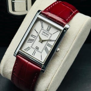 May include: A rectangular Seiko Quartz watch with a white face, black Roman numerals, and silver hands. The watch has a silver case and a red leather band. The watch is displayed on a white cushion.
