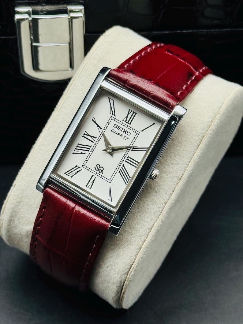 May include: A rectangular Seiko Quartz watch with a white face, black Roman numerals, and silver hands. The watch has a silver case and a red leather band. The watch is displayed on a white cushion.