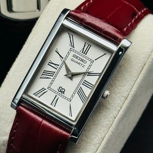 May include: A rectangular Seiko Quartz watch with a white face, black Roman numerals, and a burgundy leather band. The watch has a silver-colored case and a small crown. The band has a crocodile texture.