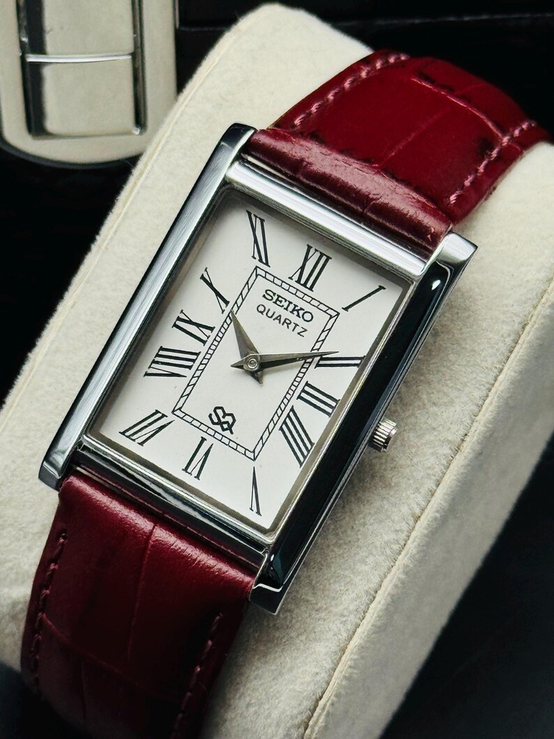 May include: A rectangular Seiko Quartz watch with a white face, black Roman numerals, and a burgundy leather band. The watch has a silver-colored case and a small crown. The band has a crocodile texture.