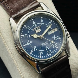 May include: A vintage Seiko 5 automatic watch with a dark blue dial and silver-tone hands and markers. The watch features a day and date display and is set on a brown leather strap. The watch face displays the text "SEIKO 5 AUTOMATIC 21 JEWELS".