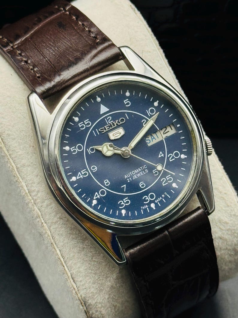 May include: A vintage Seiko 5 automatic watch with a dark blue dial and silver-tone hands and markers. The watch features a day and date display and is set on a brown leather strap. The watch face displays the text "SEIKO 5 AUTOMATIC 21 JEWELS".