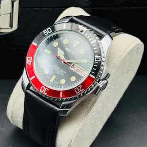 May include: A stainless steel Seiko Marine Master professional diver's watch with a black leather band. The watch features a gray face with luminous markers, a red and black rotating bezel, and a date display. The watch is displayed on a light gray cushion.