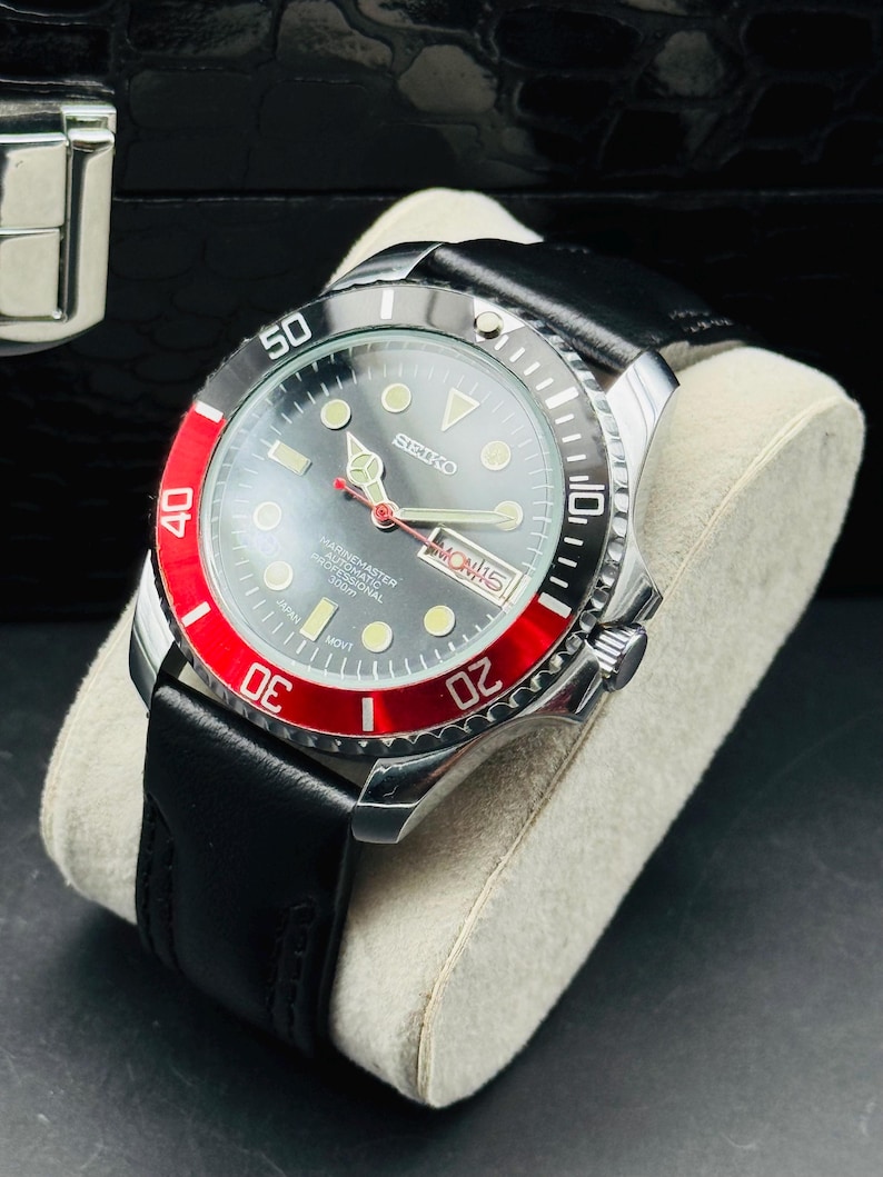 May include: A stainless steel Seiko Marine Master professional diver's watch with a black leather band. The watch features a gray face with luminous markers, a red and black rotating bezel, and a date display. The watch is displayed on a light gray cushion.