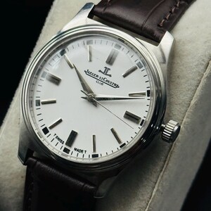 May include: A silver-toned wristwatch with a white face and black markers. The watch features the text "JAEGER-LECOULTRE CLUB" and has a brown leather band. The watch is a classic design.