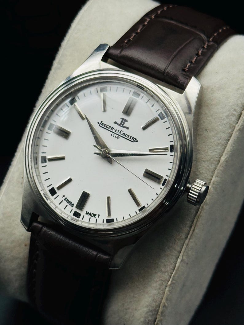 May include: A silver-toned wristwatch with a white face and black markers. The watch features the text "JAEGER-LECOULTRE CLUB" and has a brown leather band. The watch is a classic design.