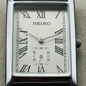 Op de afbeelding: Een rechthoekig Seiko polshorloge met een zilveren kast en een witte wijzerplaat. Het horloge heeft zwarte Romeinse cijfers, een subwijzerplaat voor seconden en een bruine leren band. De wijzers van het horloge zijn zilver.