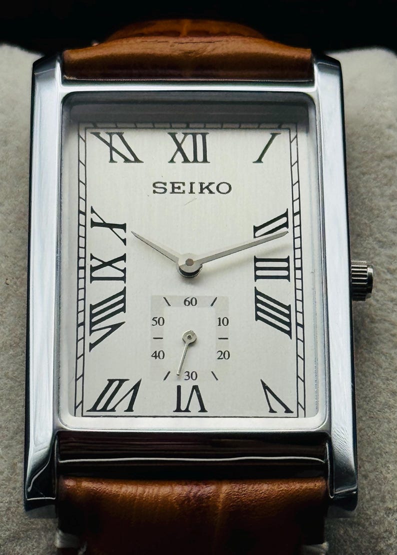 Op de afbeelding: Een rechthoekig Seiko polshorloge met een zilveren kast en een witte wijzerplaat. Het horloge heeft zwarte Romeinse cijfers, een subwijzerplaat voor seconden en een bruine leren band. De wijzers van het horloge zijn zilver.