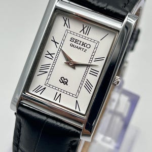 Puede incluir: Un reloj Seiko Quartz rectangular con esfera blanca, n&uacute;meros romanos negros y agujas plateadas. El reloj tiene una correa de cuero negro y una caja plateada. La esfera muestra el logotipo de Seiko y la palabra "QUARTZ".