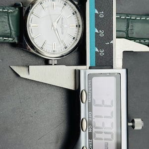 May include: A silver wristwatch with a white face and green leather strap is measured with a digital caliper. The watch face has silver markers and hands. The caliper displays a measurement of 37.30 mm.