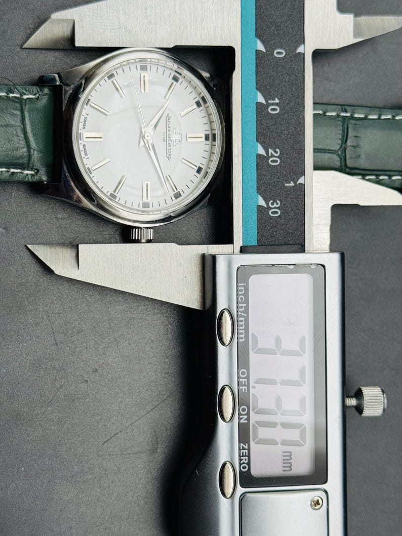 May include: A silver wristwatch with a white face and green leather strap is measured with a digital caliper. The watch face has silver markers and hands. The caliper displays a measurement of 37.30 mm.