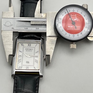 Puede incluir: Un reloj Seiko Quartz plateado con esfera rectangular y correa de cuero negro se mide con un calibrador. La esfera del reloj tiene n&uacute;meros romanos y el nombre de la marca. El calibrador muestra una medida.