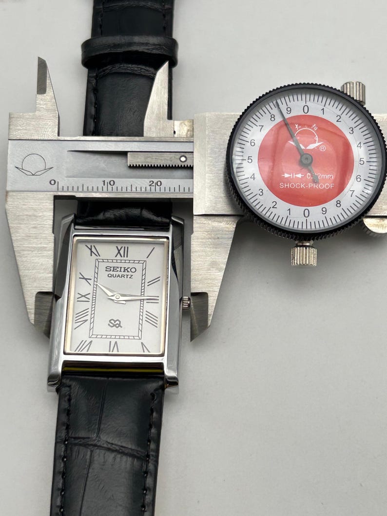 Puede incluir: Un reloj Seiko Quartz plateado con esfera rectangular y correa de cuero negro se mide con un calibrador. La esfera del reloj tiene n&uacute;meros romanos y el nombre de la marca. El calibrador muestra una medida.