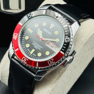 May include: A Seiko Marine Master Professional 300m watch with a black dial, red bezel, and black leather strap. The watch features luminous markers, a date display, and a stainless steel case. The watch is displayed on a gray cushion.