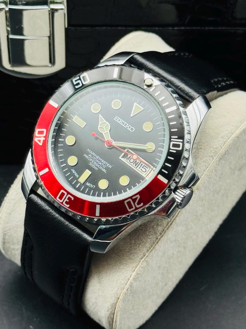 May include: A Seiko Marine Master Professional 300m watch with a black dial, red bezel, and black leather strap. The watch features luminous markers, a date display, and a stainless steel case. The watch is displayed on a gray cushion.
