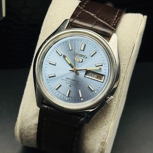 May include: A silver-toned Seiko 5 Sports watch with a light blue dial and a brown leather strap. The watch face displays the day and date. The watch is displayed on a cream-colored cushion.