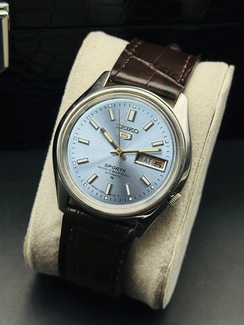 May include: A silver-toned Seiko 5 Sports watch with a light blue dial and a brown leather strap. The watch face displays the day and date. The watch is displayed on a cream-colored cushion.