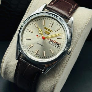 May include: A silver Seiko automatic wristwatch with a light silver dial, gold hands, and a red second hand. The watch has a brown leather strap and displays "TUE 23". The watch is displayed on a white cushion.