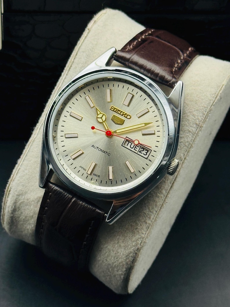May include: A silver Seiko automatic wristwatch with a light silver dial, gold hands, and a red second hand. The watch has a brown leather strap and displays "TUE 23". The watch is displayed on a white cushion.