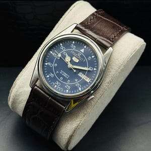 May include: A silver-toned Seiko automatic watch with a dark blue face and brown leather strap. The watch face displays the day and date, with white numerals and hands. The watch is displayed on a cream-colored cushion.